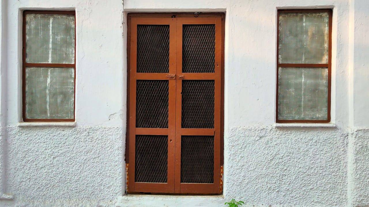 A rustic wooden door with iron mesh flanked by two windows on a white-textured wall.