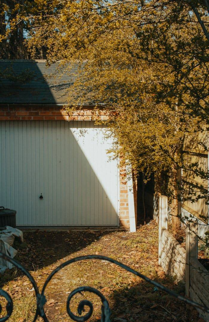 A sunlit garden in England with a white garage door surrounded by foliage.