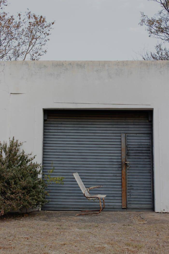 An abandoned building with a roller door and a lone chair outside on a dry landscape.