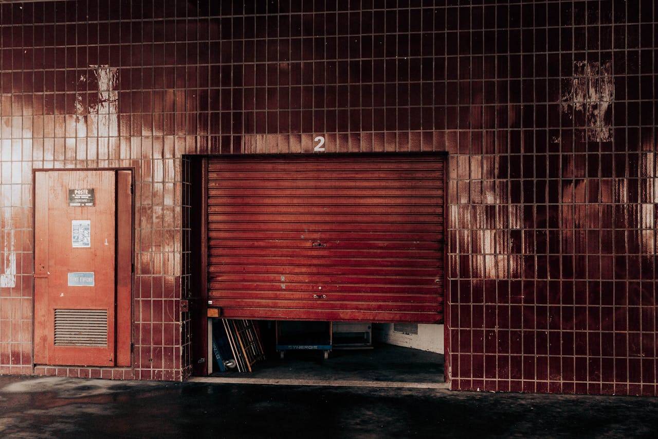 Aged garage exterior with security shutter and door between tiled wall with number on sunny day