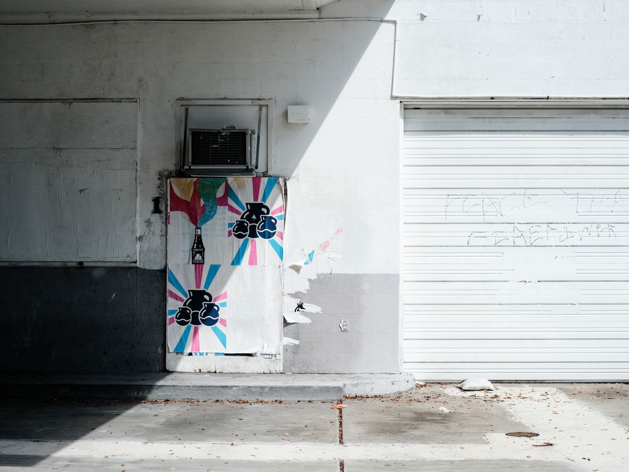 Street art on a sunlit wall with a white garage door and colorful posters.