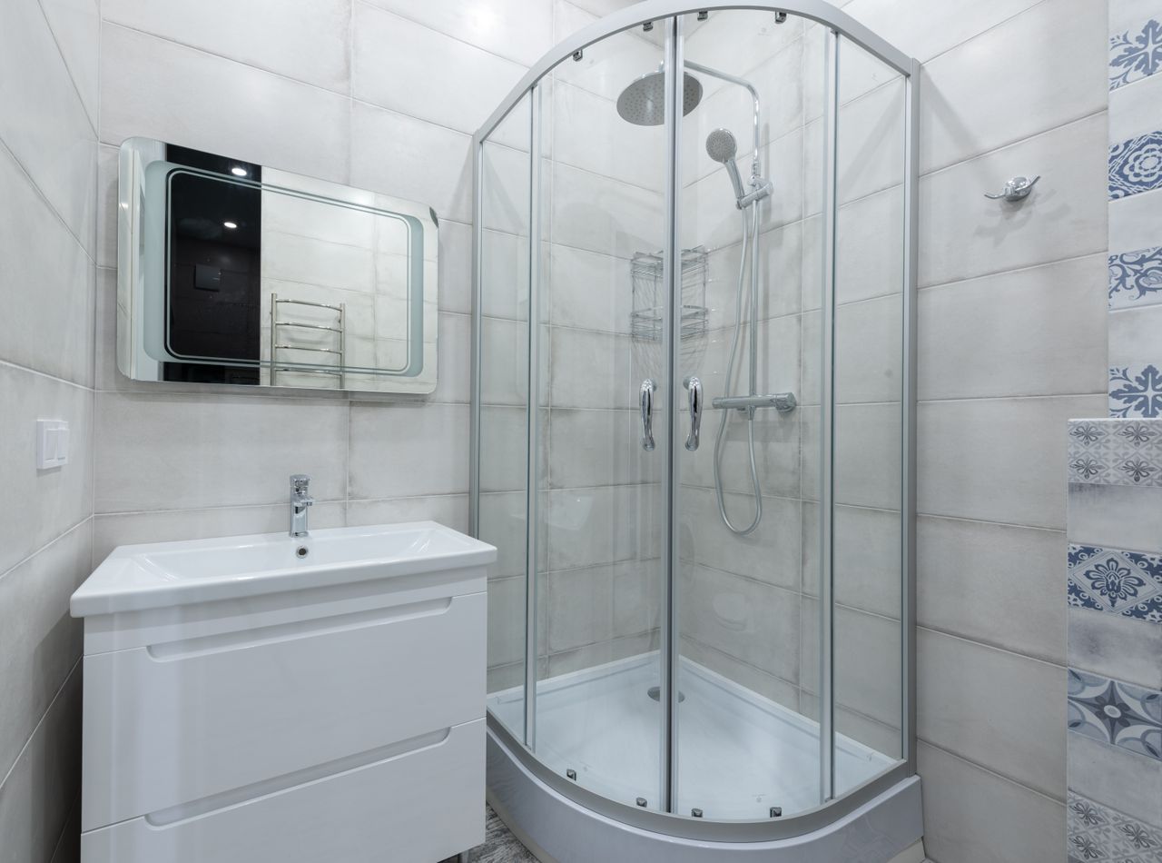 Interior of contemporary light washroom with modern glass shower cabin placed near wall with white sink and mirror at home