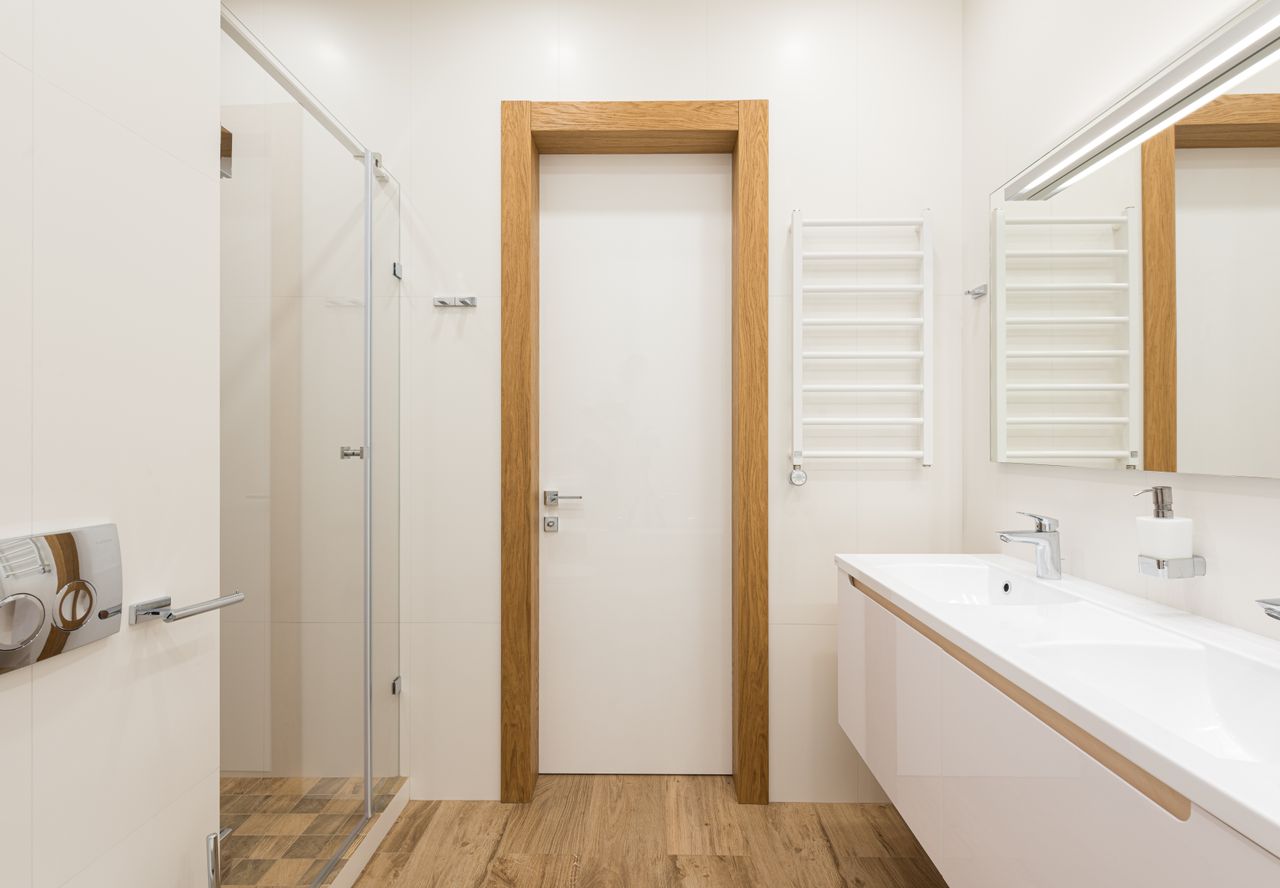 White door and furniture in modern bathroom with glass shower cabin and wooden elements reflecting in mirror