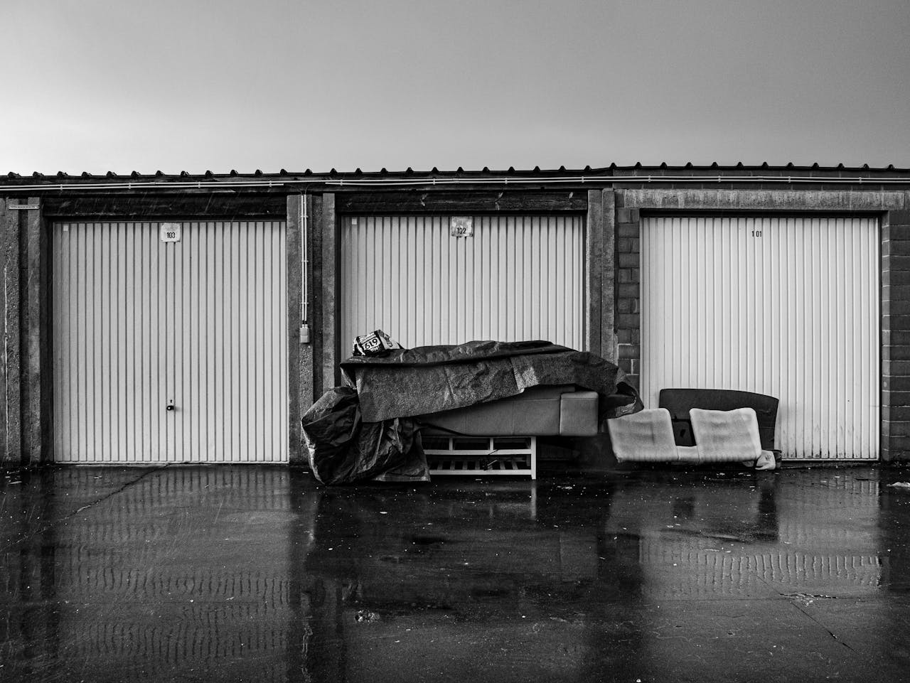 Monochrome view of closed storage units with covered furniture outdoors on a rainy day.